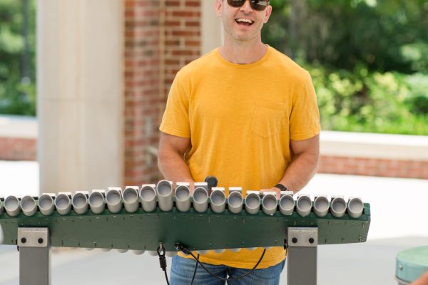 Man in yellow shirt playing outdoor percussion instrument with metal tubes on green frame in park setting