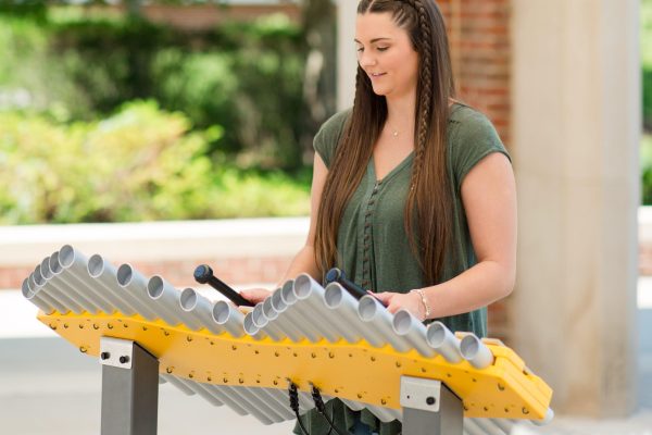 Woman interacting with yellow outdoor musical instrument featuring metal tubes, designed for creative park music play and collaboration.