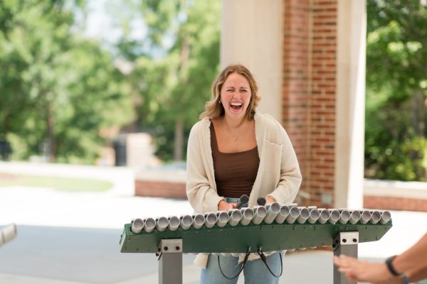 Woman playing outdoor metal tube musical instrument surrounded by greenery and brick columns in a park setting