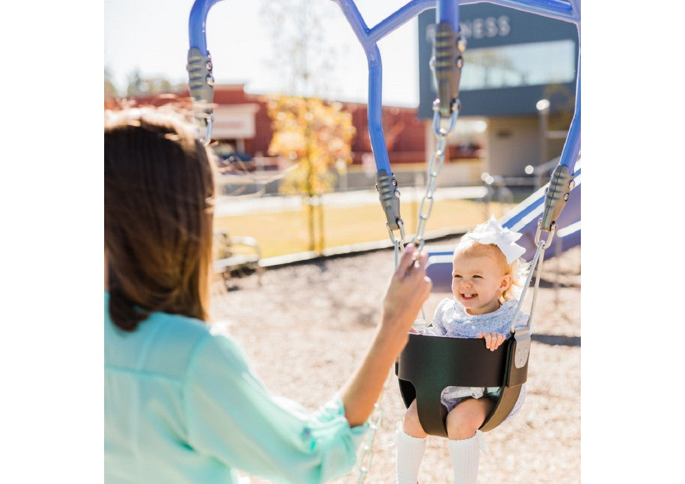 Toddler with white bow seated in black bucket swing held by adult in light blue shirt at playground