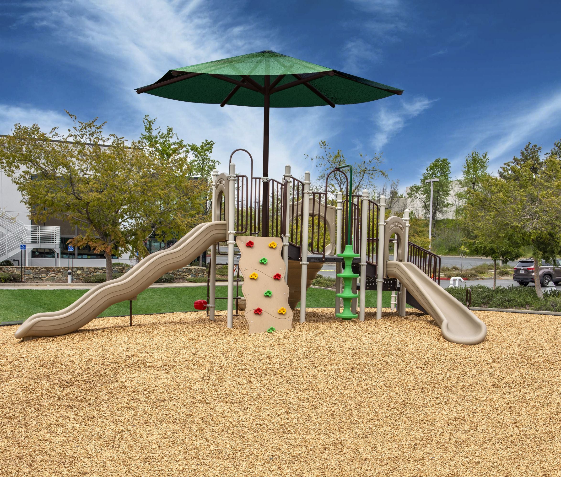 Outdoor playground structure with four slides, climbing wall, overhead ladder, green shade canopy, and wood chip ground cover