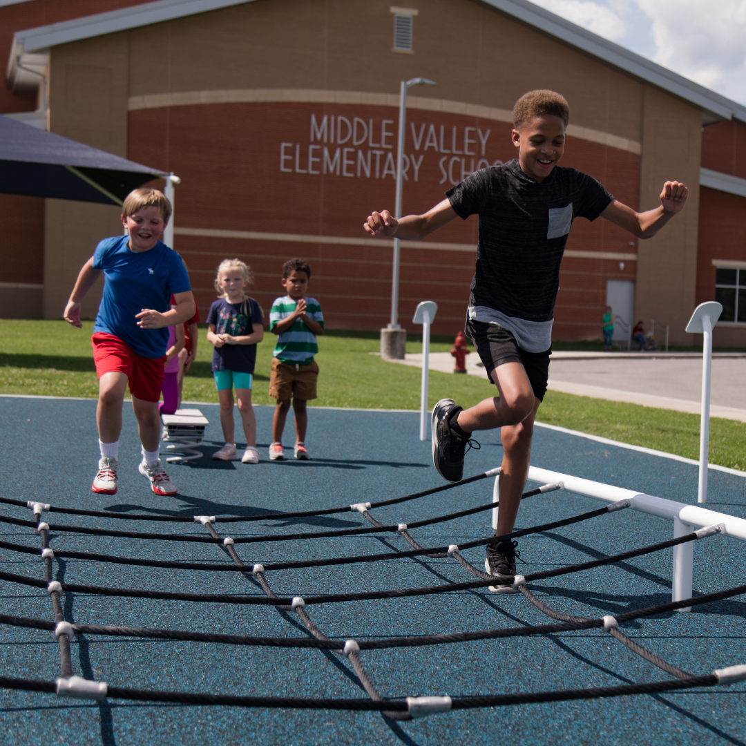 Children climbing and racing on a black rope net structure with blue safety surfacing at an elementary school playground