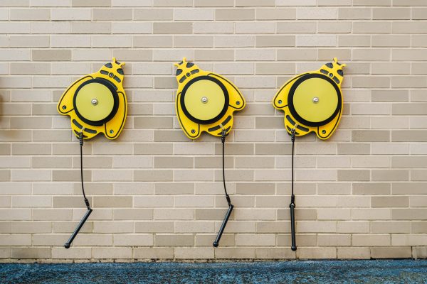 Three yellow bee-shaped outdoor musical instruments with attached mallets mounted on a brick wall for interactive play