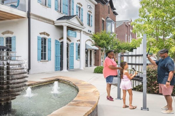Outdoor musical instrument with vertical anodized aluminum tubes, played by diverse people near a water fountain and residential buildings.