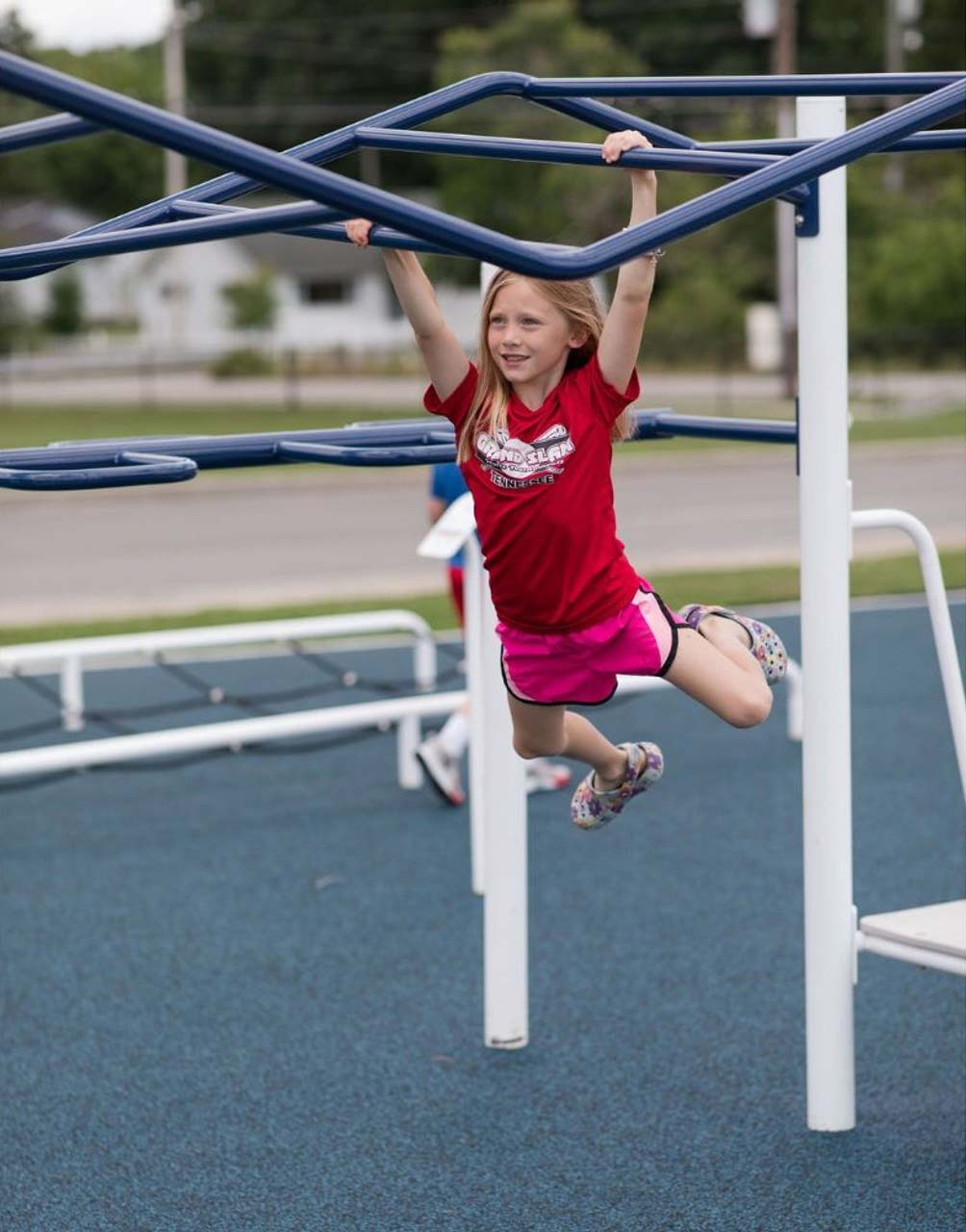 Child in red shirt and pink shorts using blue accessible outdoor strength bars for kids' fitness and coordination