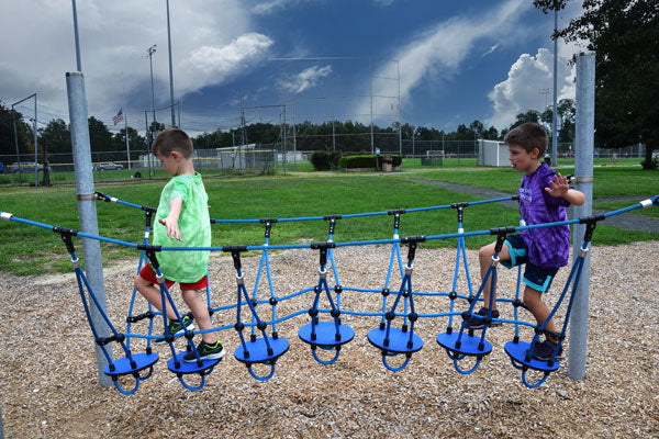 Two children balancing on a blue wobble disk adventure bridge with galvanized steel posts in an outdoor playground