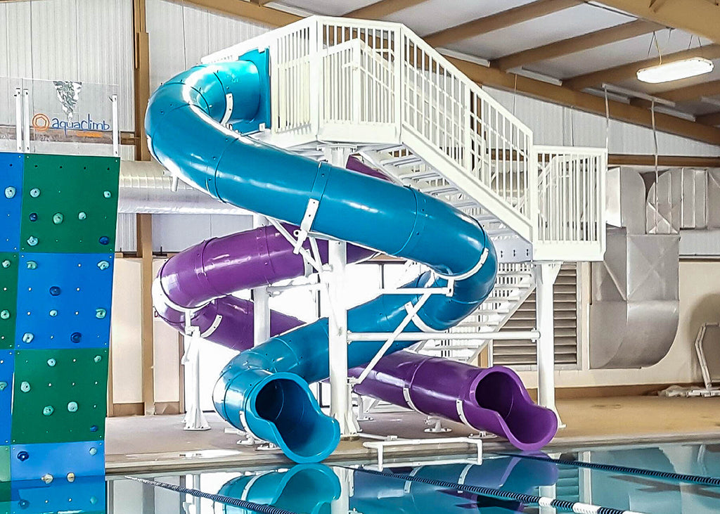 Dual twisting blue and purple water slides with open white stair access beside an indoor pool and climbing wall