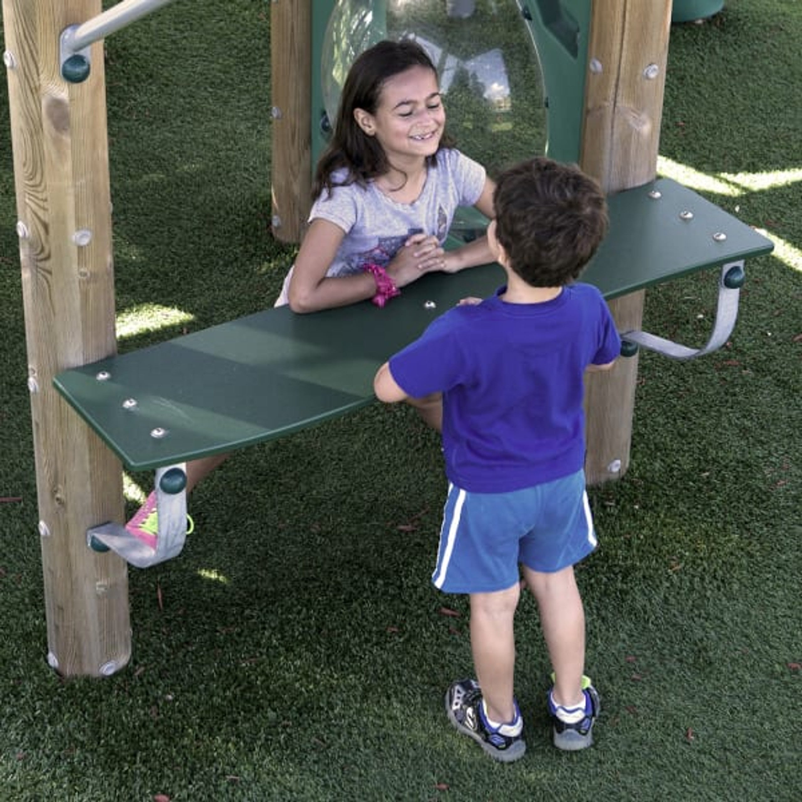 Two children at an ADA-accessible green play table with wooden posts on artificial turf, featuring interactive play elements.