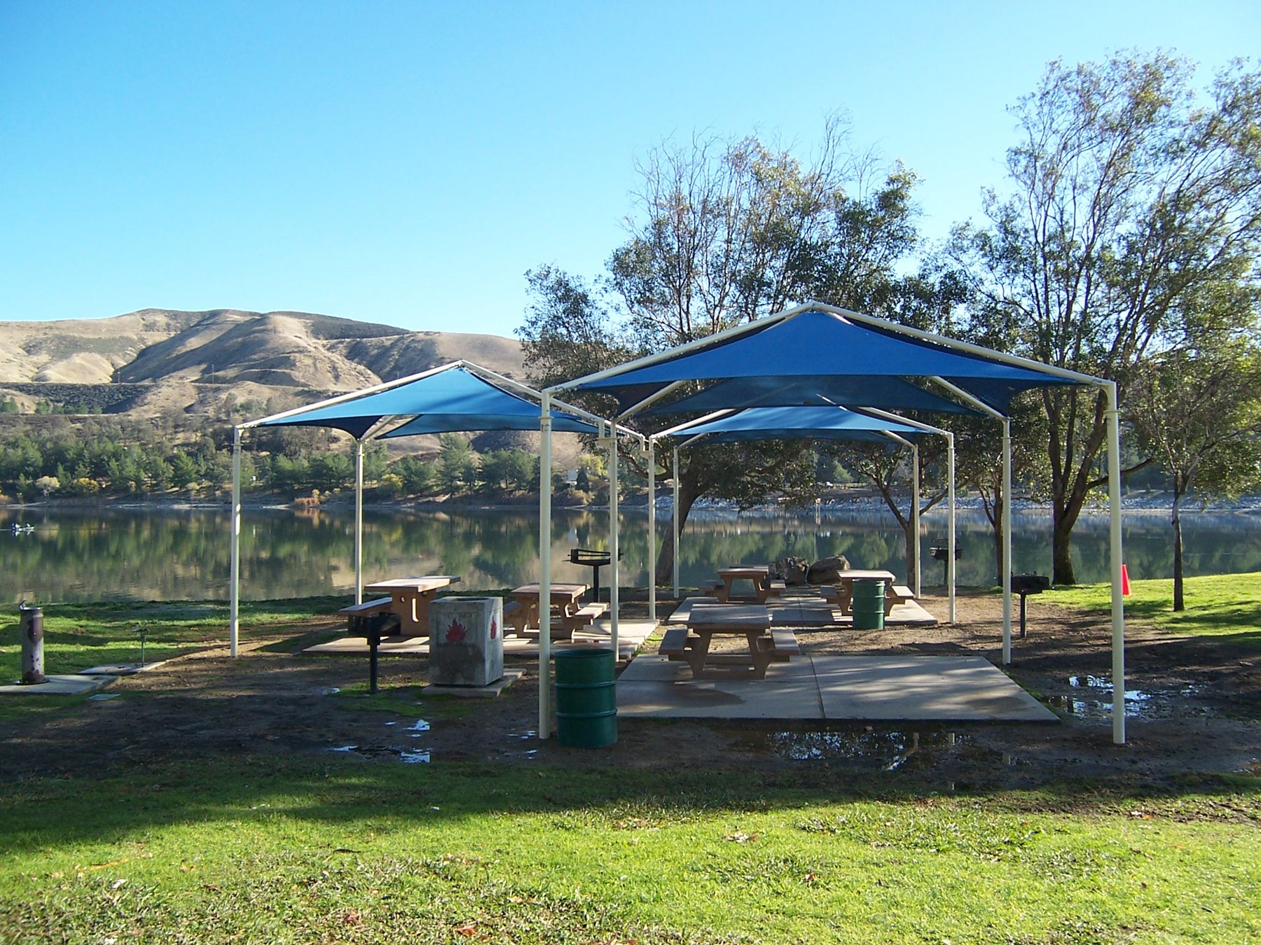Pyramid Multi-Panel Shade Structure with four blue fabric panels over picnic tables at lakeside park
