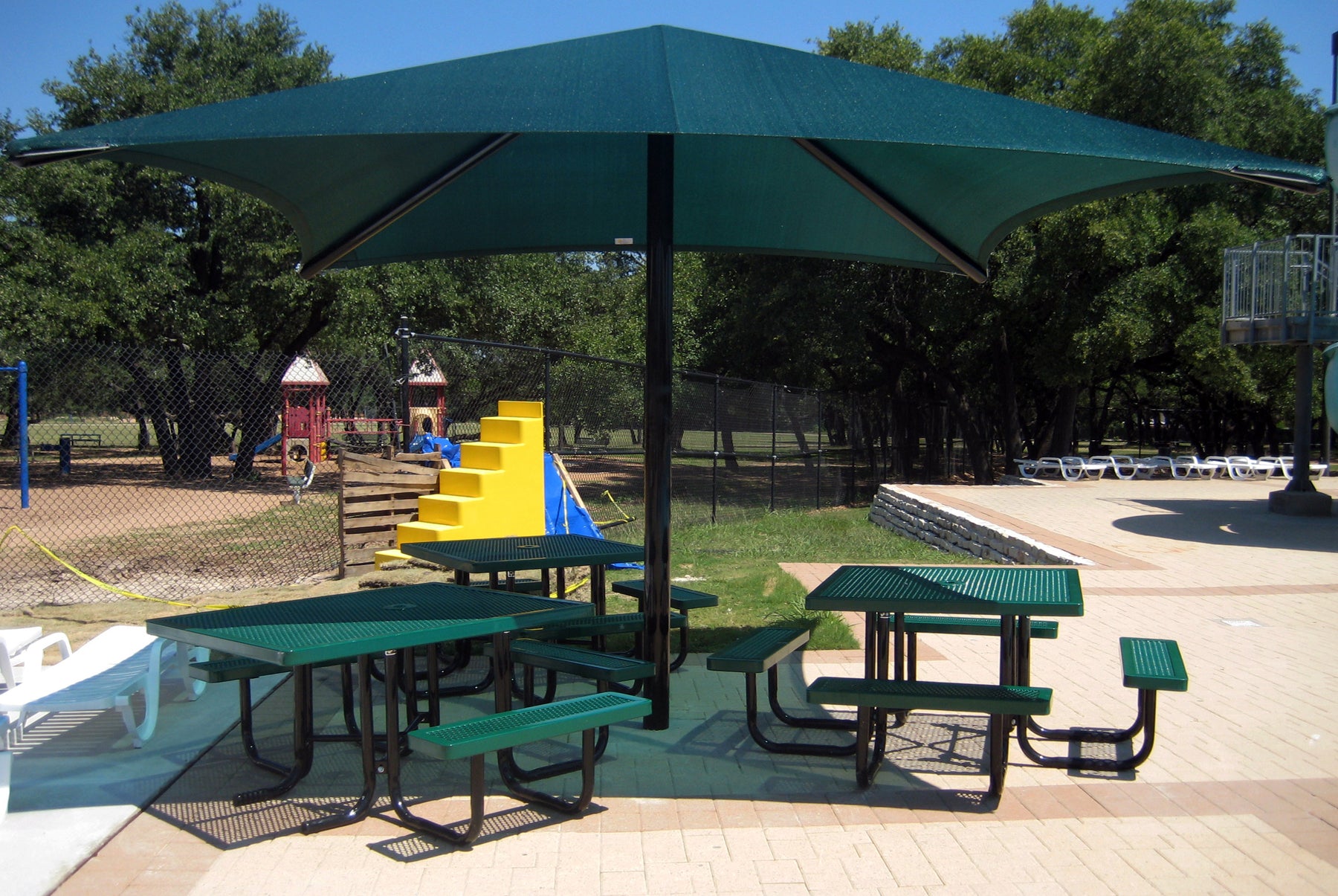 Green single post pyramid shade structure over picnic tables near playground and pool area, providing UV protection