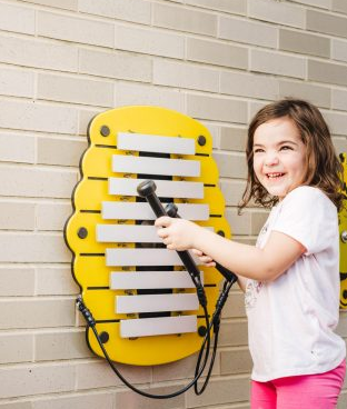 Child playing a yellow beehive-shaped outdoor metallophone with tethered mallets mounted on a brick wall