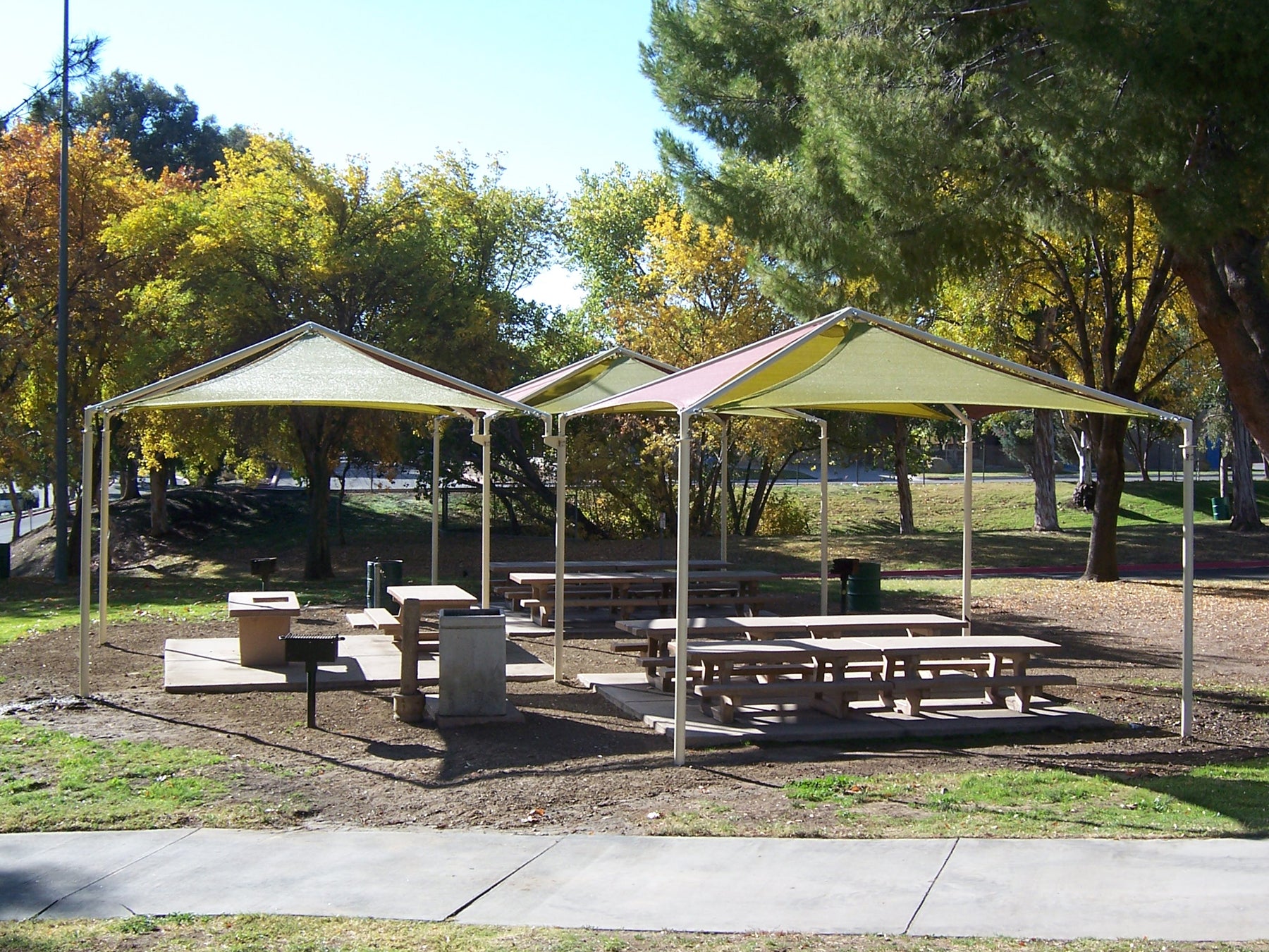 Pyramid shade structure with four green and pink fabric panels covering picnic tables in a park setting
