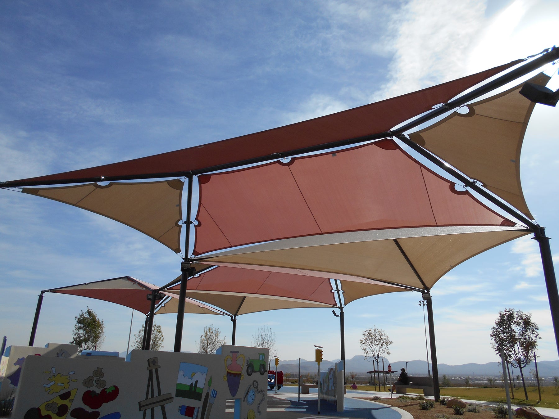 Outdoor playground shade with four muted red and beige fabric panels supported by black metal posts.