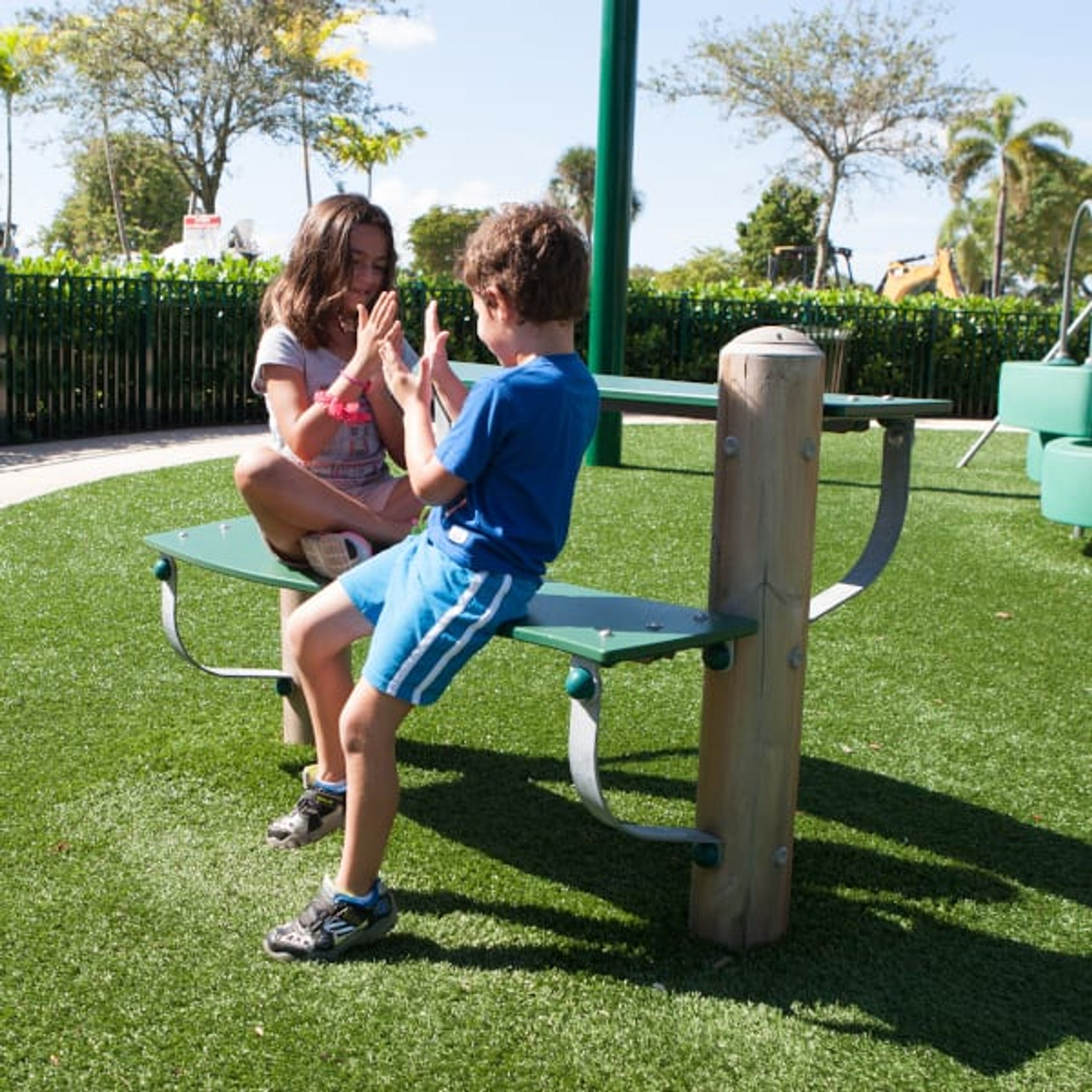 Two children playing hand-clap game on green outdoor play bench on artificial turf in playground setting
