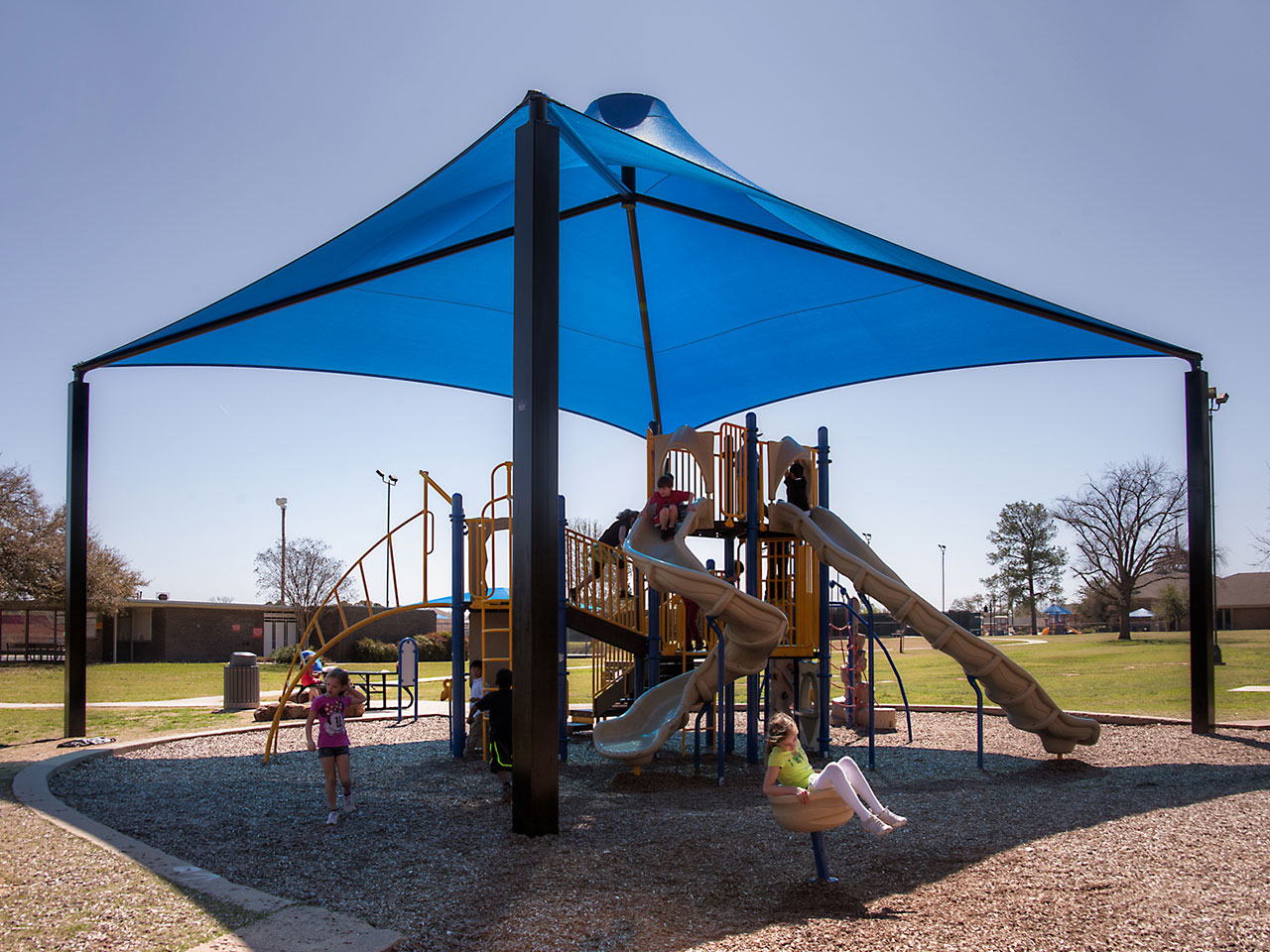 Blue conical shade structure with four black posts covering outdoor playground slides and climbing equipment