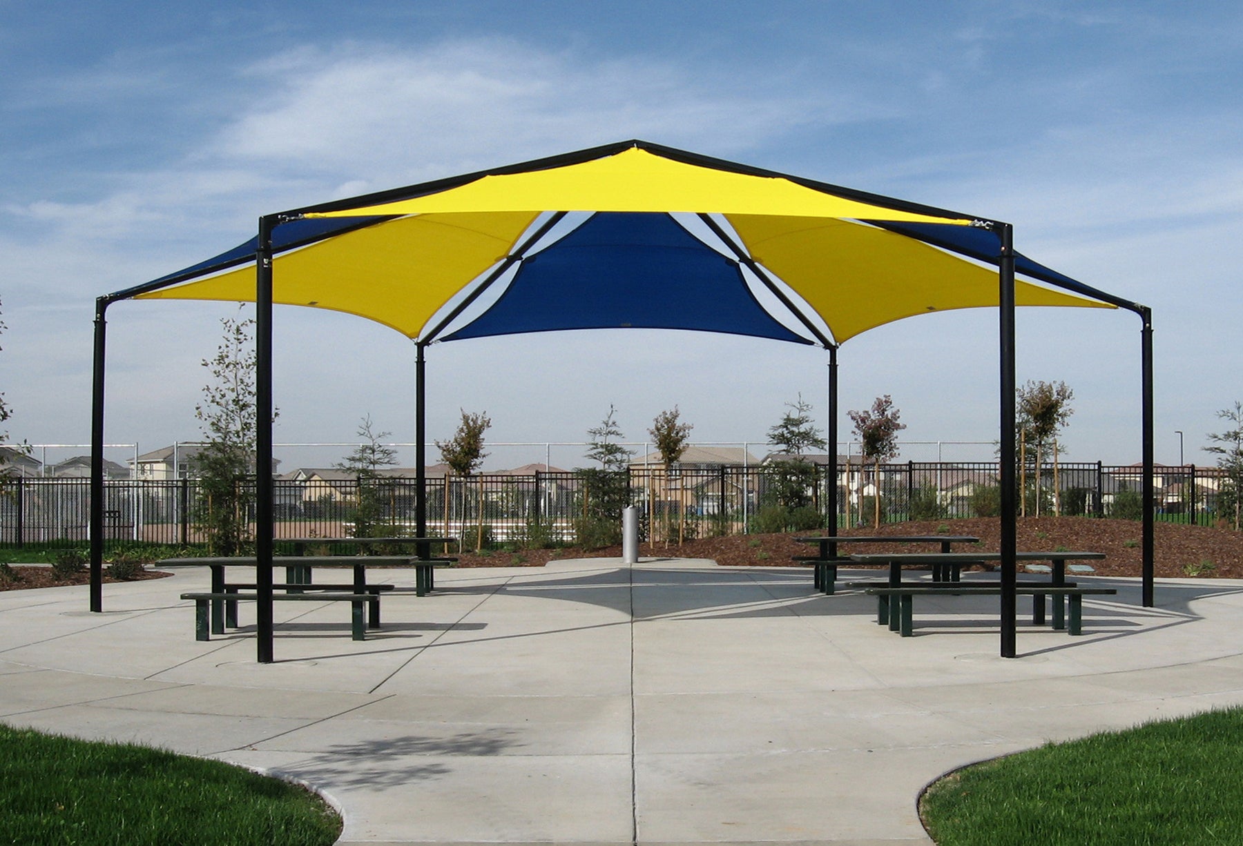 Hexagonal outdoor shade structure with alternating yellow and blue fabric panels over picnic tables in a park setting