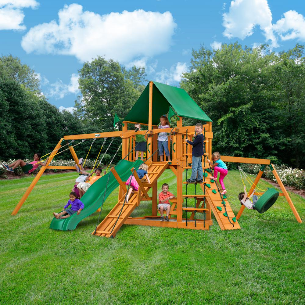 Wooden playset with green vinyl canopy, multiple swings, climbing ramp, slide, and children playing on grass outdoors