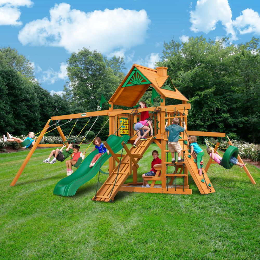 Cedar wooden playset with green slide, two belt swings, climbing ramp, tire swing, and roofed play deck with children playing