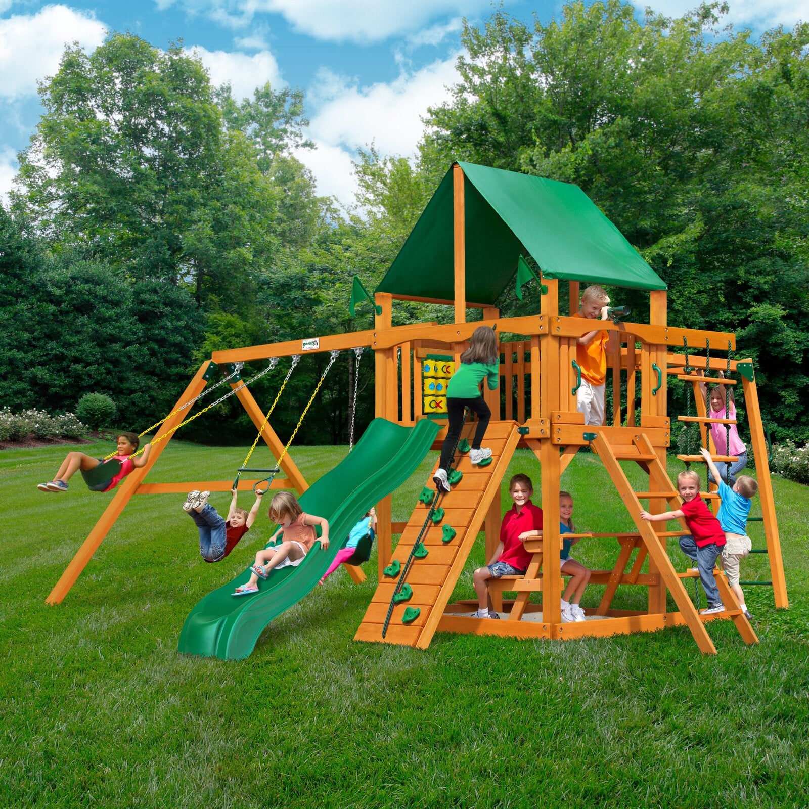 Cedar wooden playset with green vinyl canopy, slide, swings, climbing wall, monkey bars, picnic table, and sandbox area outdoors