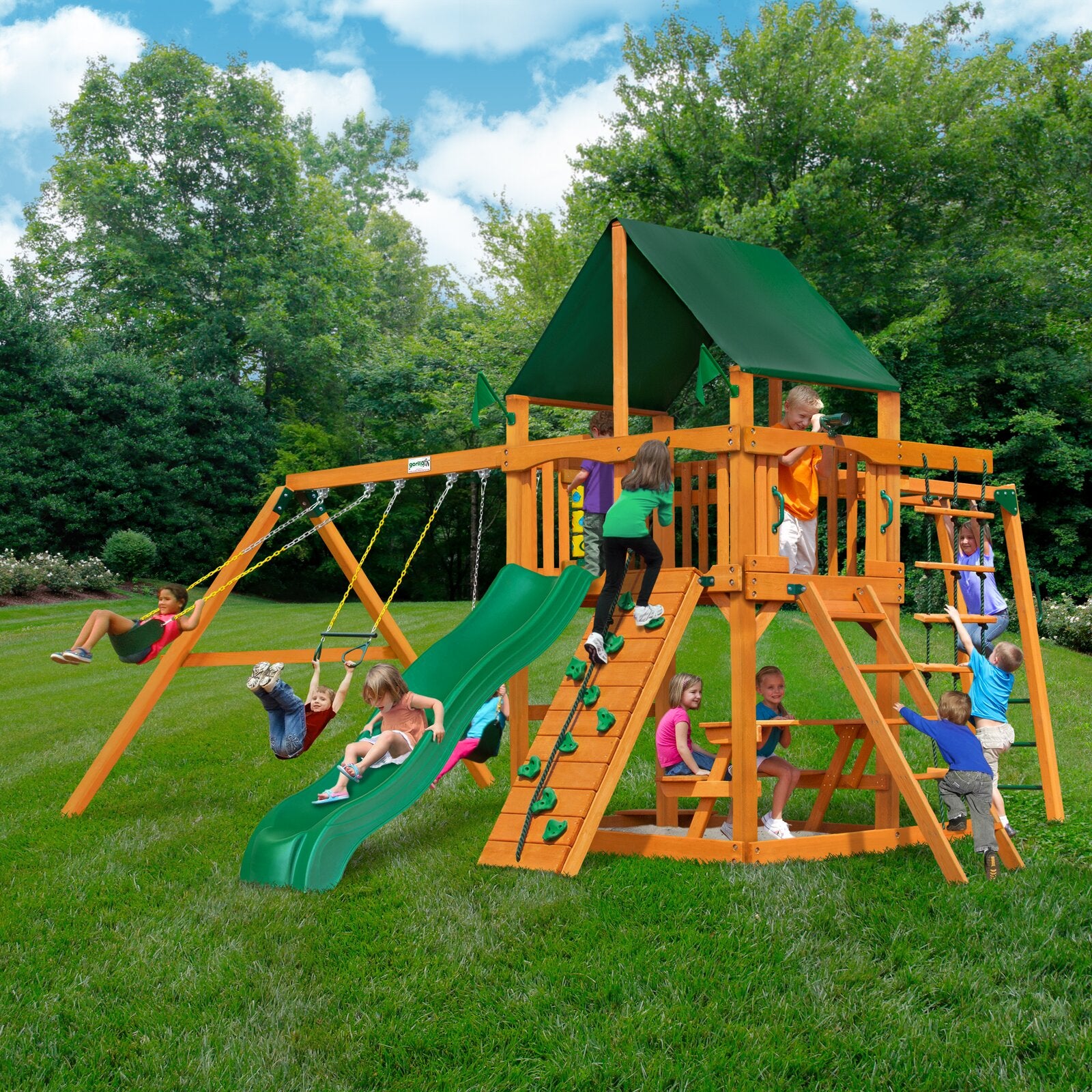 Wooden playset with green vinyl canopy, slide, swings, climbing wall, rope ladder, monkey bars, picnic table, and sandbox with children playing