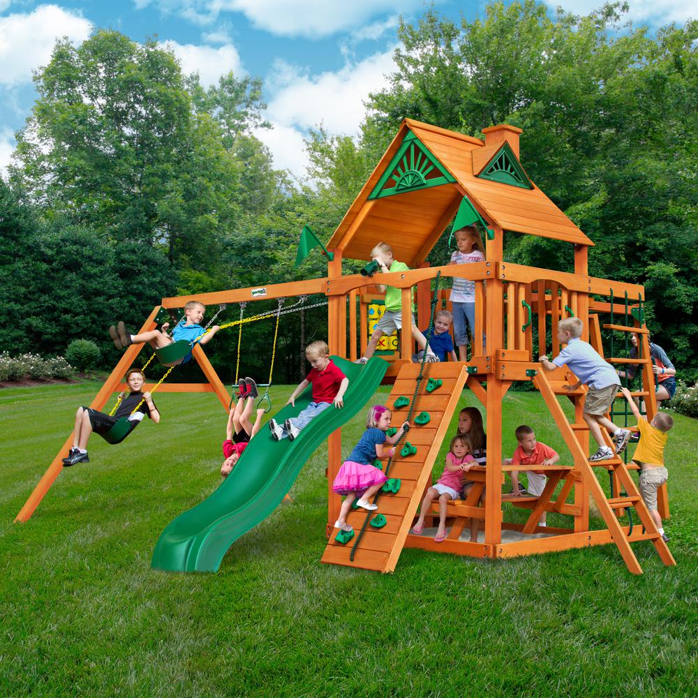 Cedar wooden swing set with green slide, rock climbing wall, trapeze bar, swings, picnic table, and sandbox with children playing outdoors