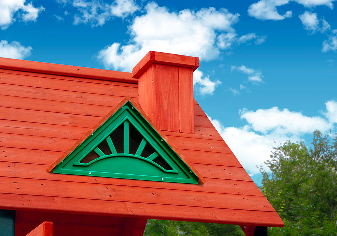 Redwood-stained wooden playset roof with green sunburst dormer, chimney, and blue sky with clouds and trees.