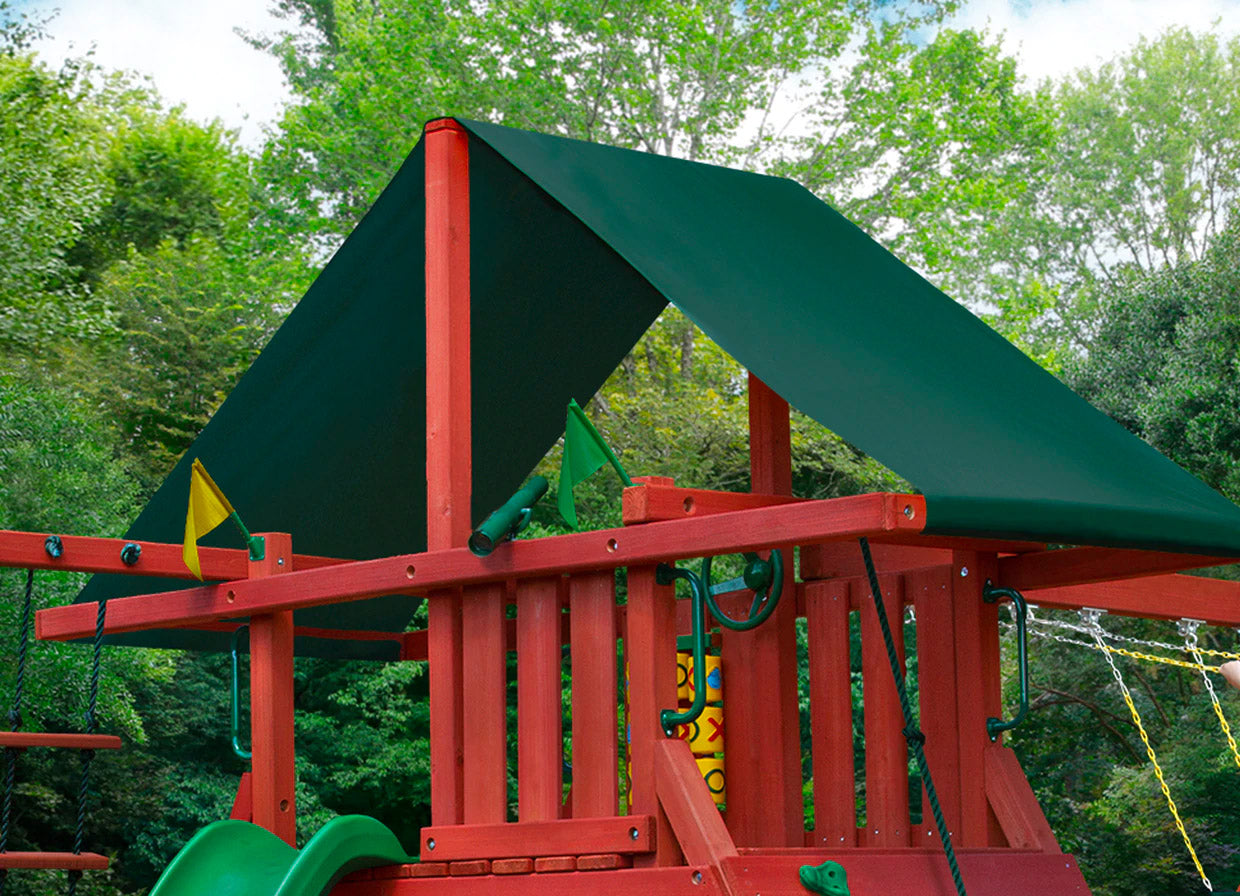 Redwood-finished wooden swing set with green Sunbrella canopy, rock wall, rope ladder, tic-tac-toe panel, and multiple swings in a backyard setting