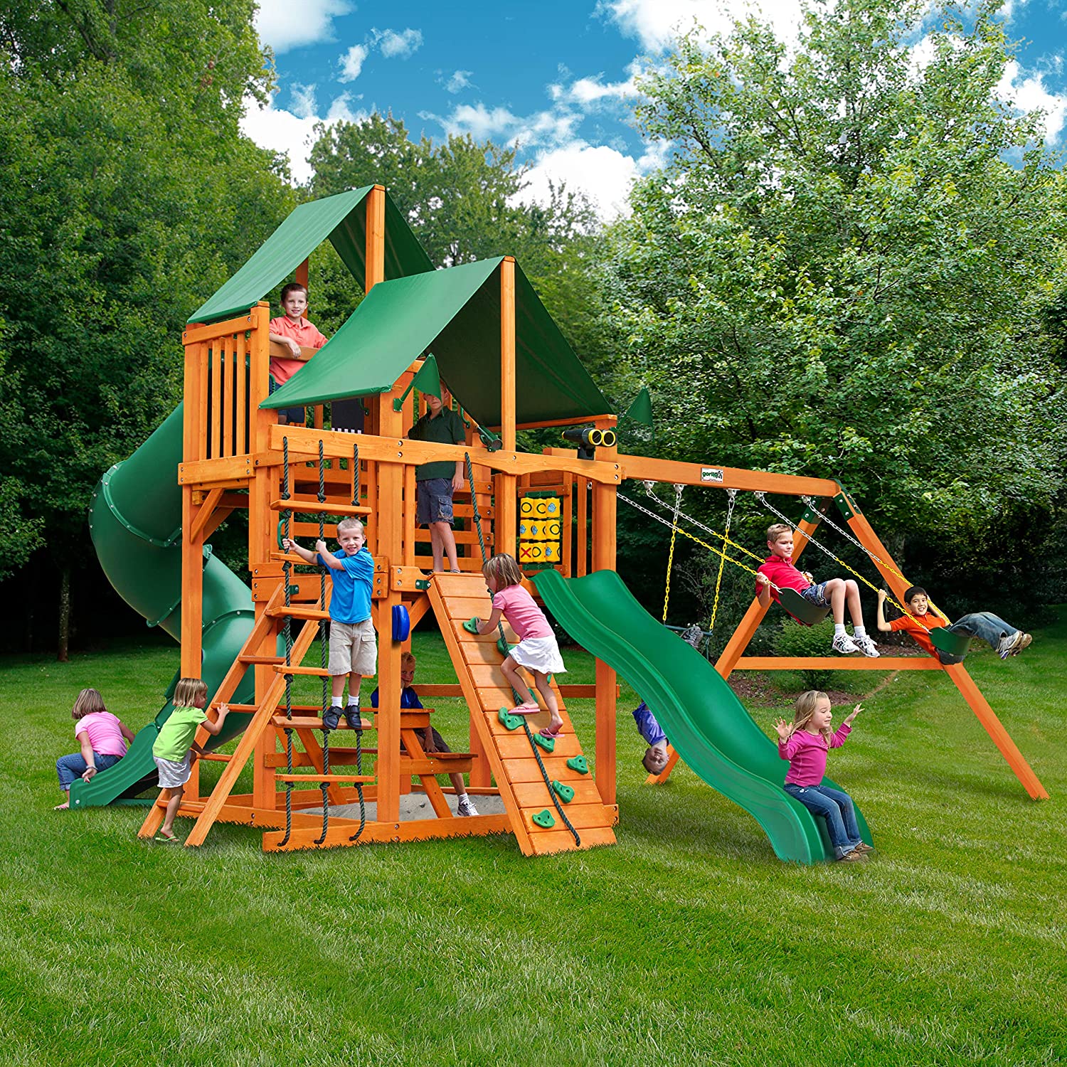 Wooden playset with green vinyl canopy, dual belt swings, turbo tube slide, climbing wall, rope ladder, tic-tac-toe panel, and sandbox with children playing