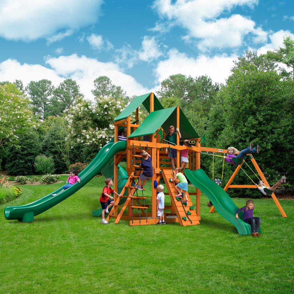 Wooden playset with green vinyl canopy, two slides, climbing wall, rope ladder, swings, and children playing on grass