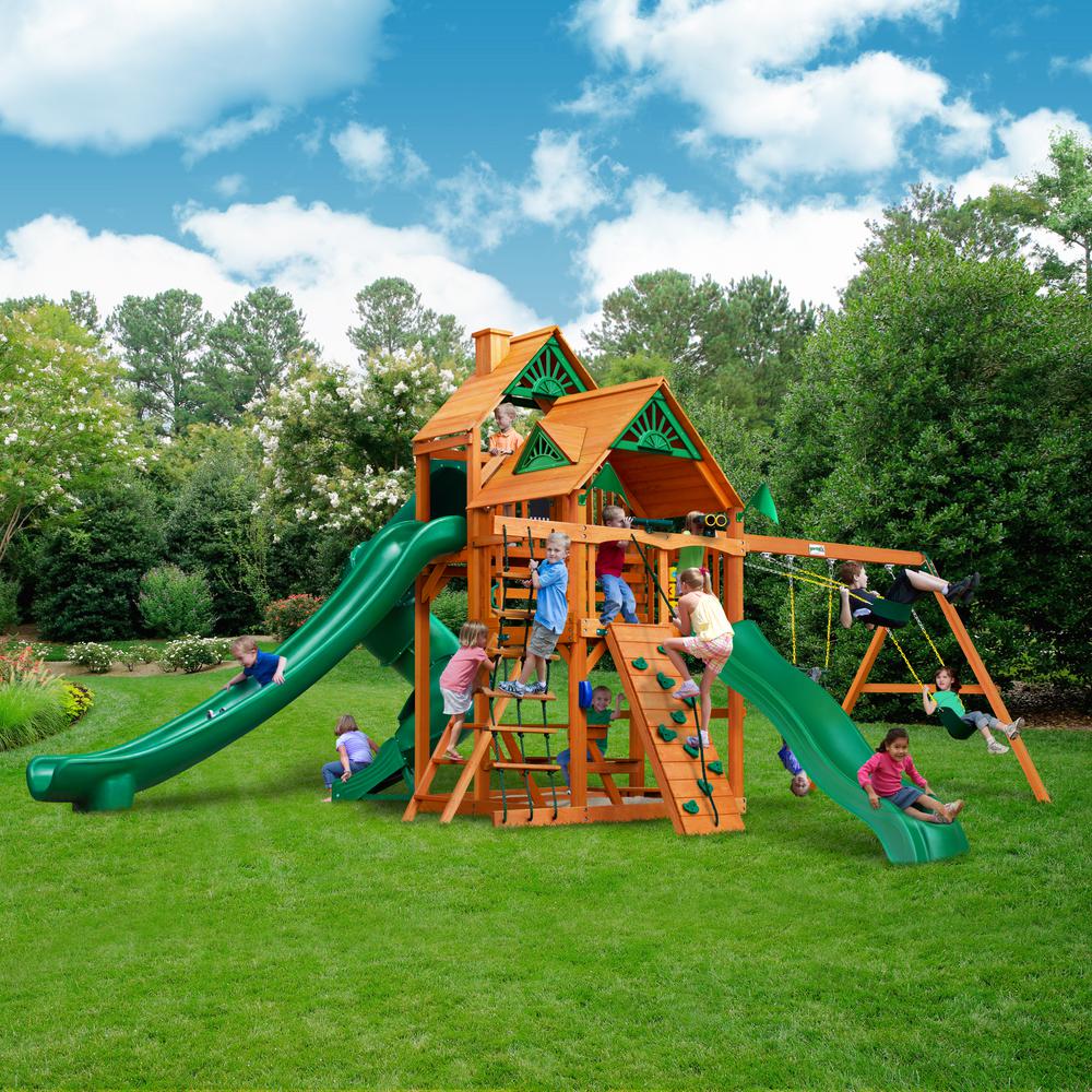 Cedar wooden playset with two green slides, climbing wall, rope ladder, swings, and playhouse roof outdoors