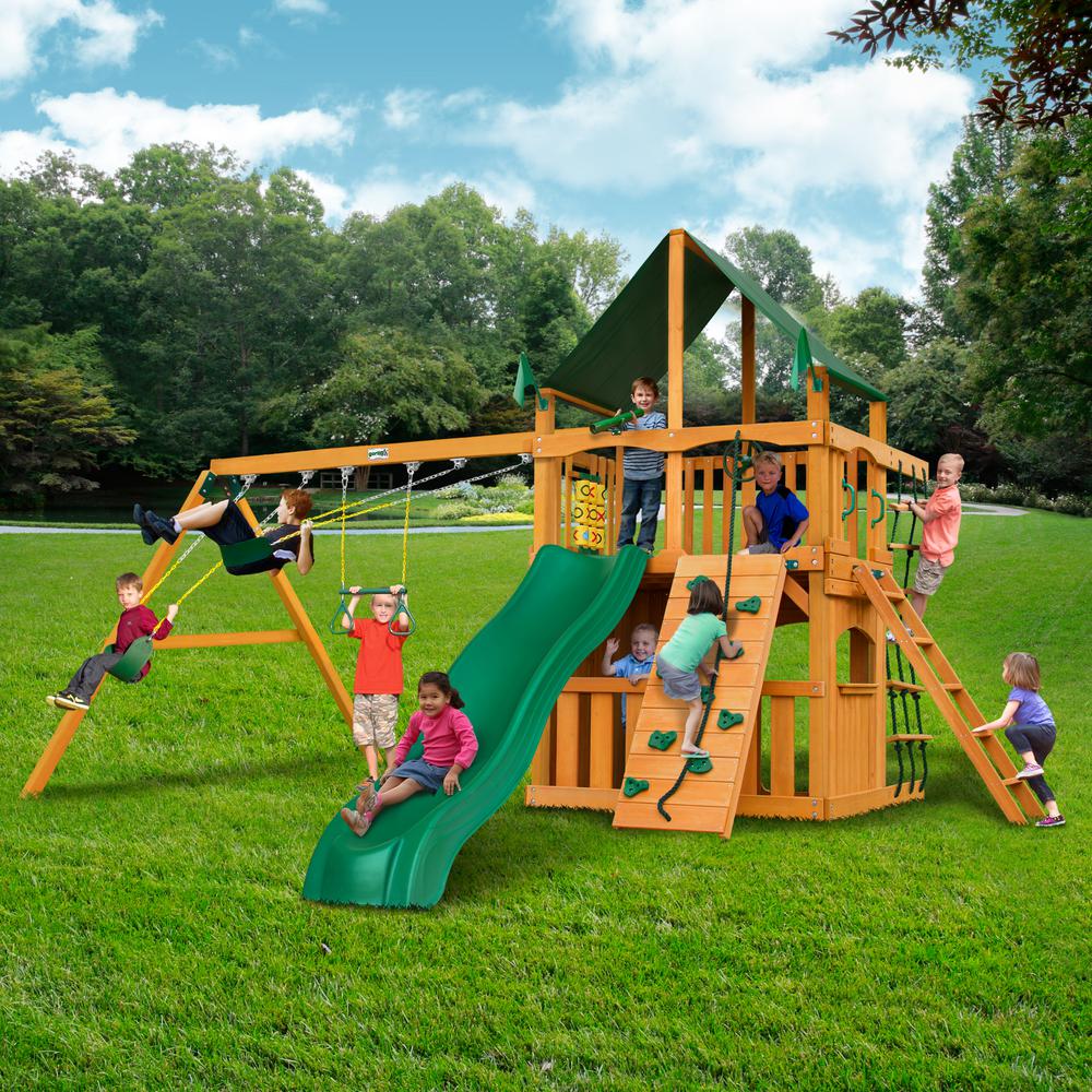 Cedar wooden swing set with green slide, two belt swings, trapeze bar, climbing walls, rope ladder, and tic-tac-toe panel outdoors with children playing