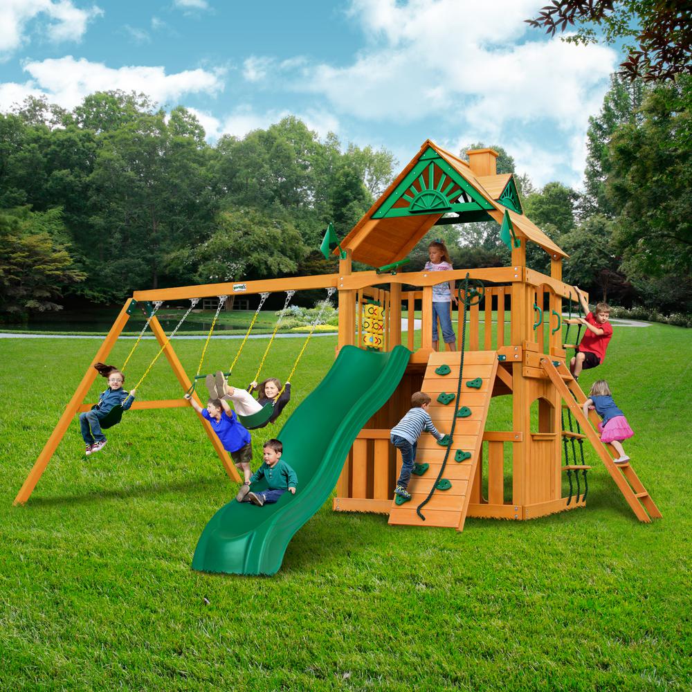 Cedar wooden swing set with green slide, rock climbing wall, rope ladder, tic-tac-toe panel, and children playing outdoors