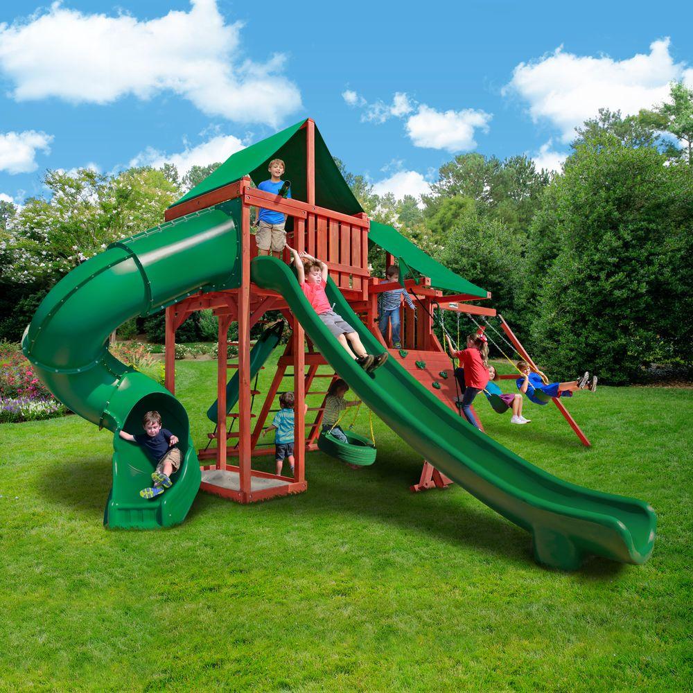 Redwood-finished wooden playset with green canopy, two green slides, tire swing, belt swings, climbing wall, and children playing outdoors