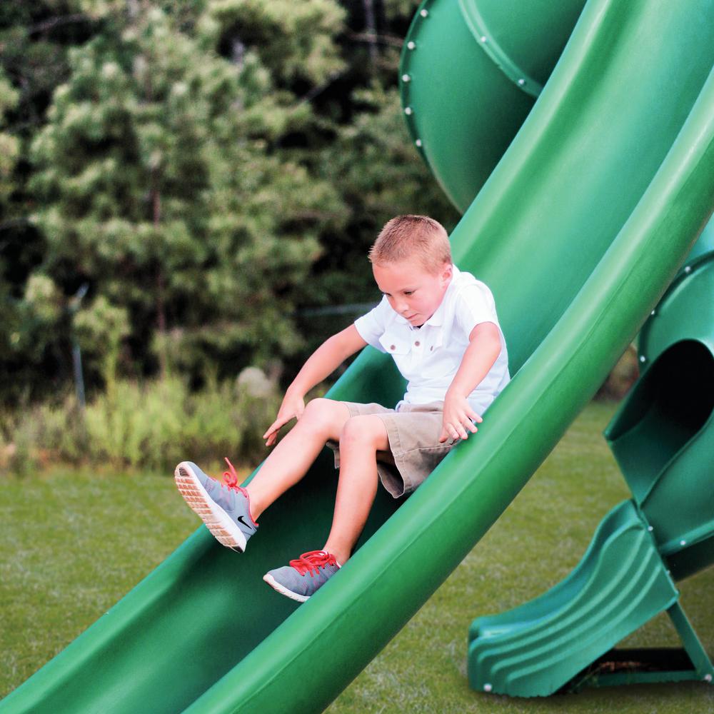 Young boy sliding down a green curved plastic slide on a wooden swing set in a grassy backyard with play features.