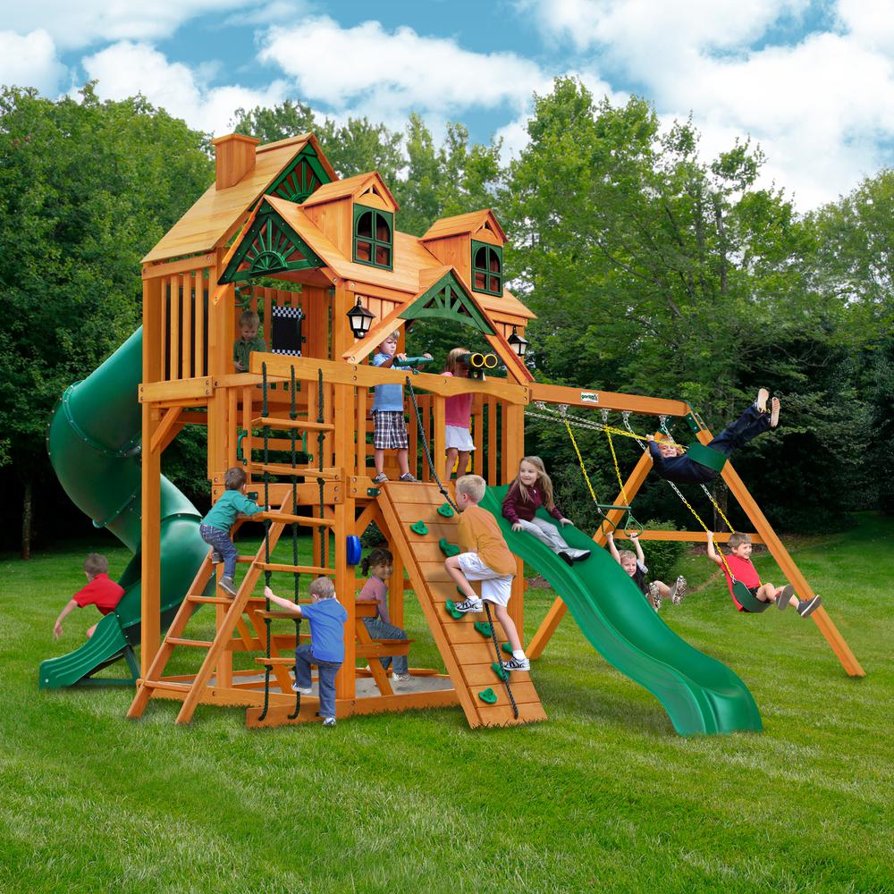 Wooden playset with amber finish, green slide, dual swings, climbing wall, rope ladder, and Malibu wood roof with dormers and chimney