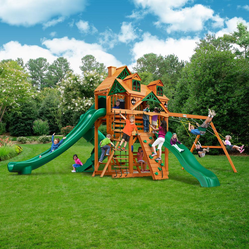 Cedar wooden playset with green slides, climbing wall, swings, playhouse roof with decorative windows and chimney, solar wall lights.