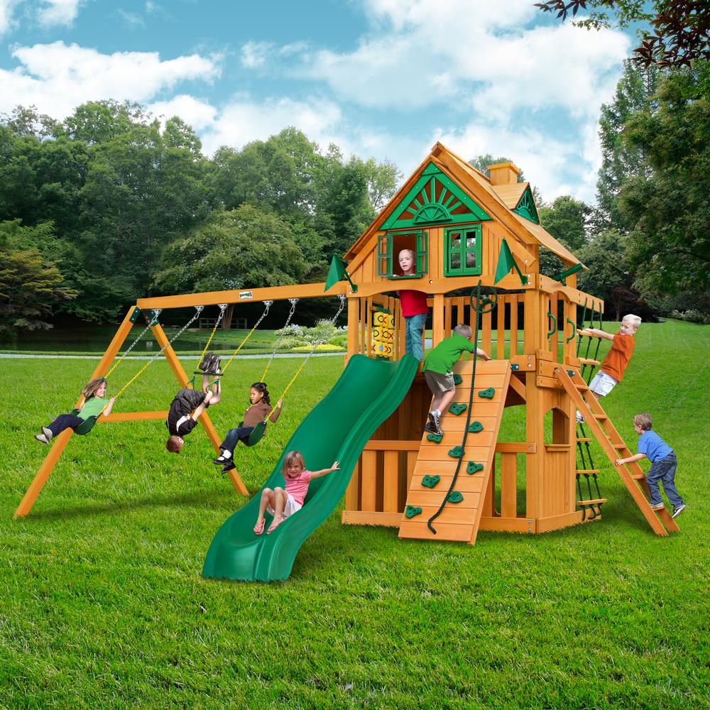 Cedar wooden playset with green slide, two belt swings, trapeze swing, climbing wall, rope ladder, and upper clubhouse with windows and shutters