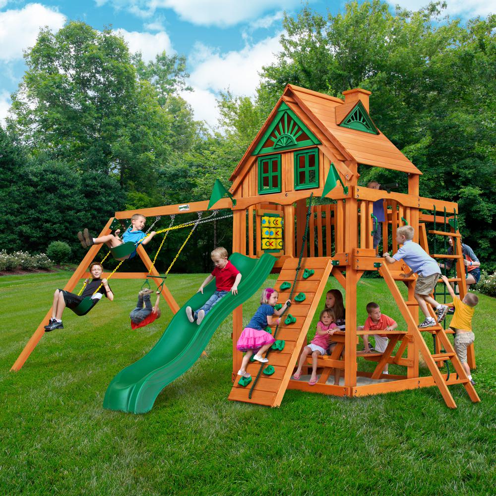Cedar wooden playset with green slide, two belt swings, rock climbing wall, picnic table, and tic-tac-toe panel in backyard setting