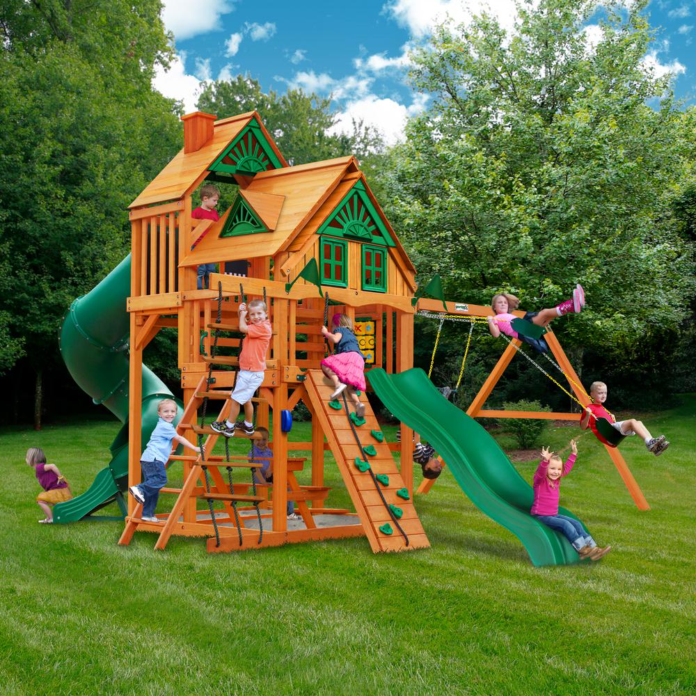 Cedar wooden playset with green slides, climbing wall, rope ladder, dual swings, trapeze, and sunburst-detailed wood roof