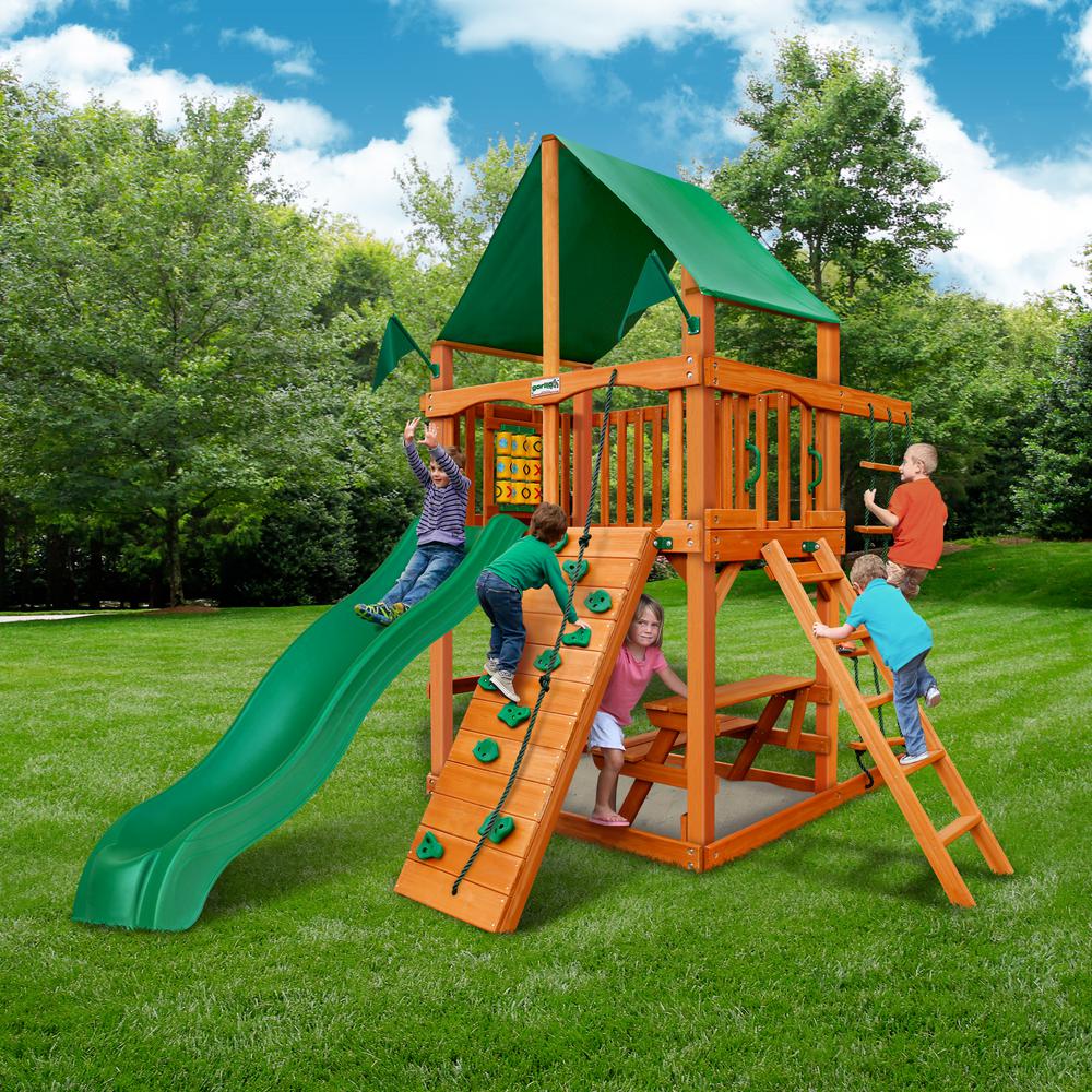 Wooden playset with green vinyl canopy, rock climbing wall, slide, ladder, and tic-tac-toe panel in grassy backyard