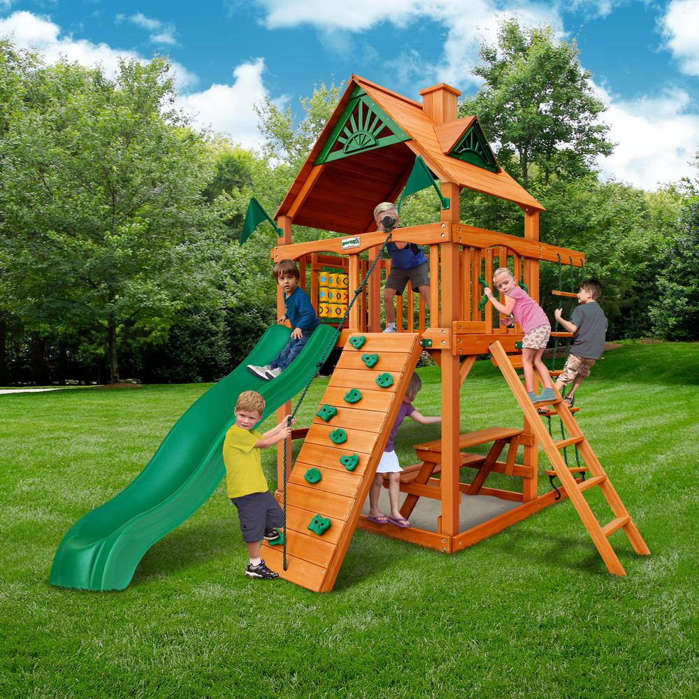 Wooden playset with green slide, rock climbing wall, rope ladder, tic-tac-toe panel, and picnic table with children playing