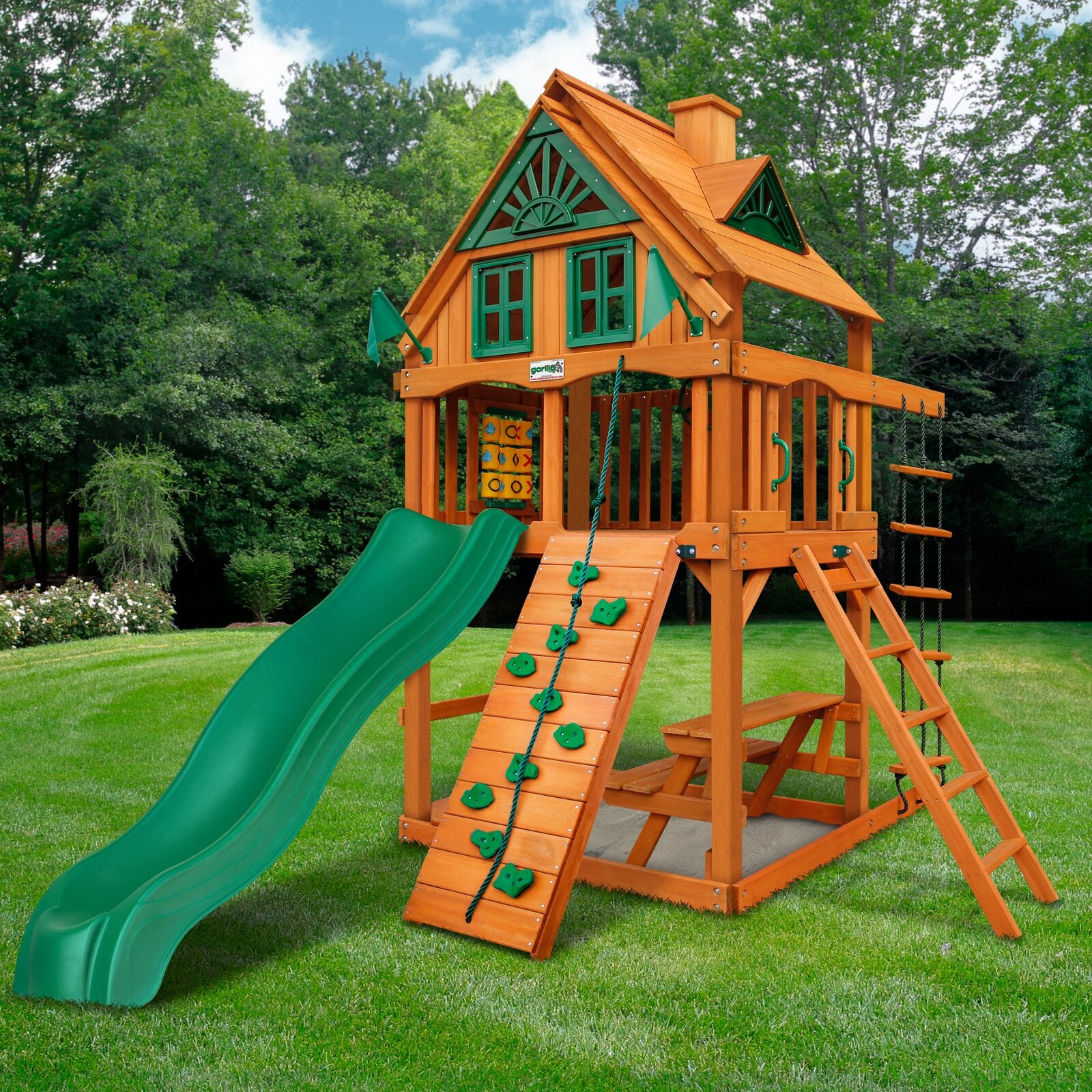 Cedar wooden playset with green wave slide, rock climbing wall, rope ladder, tic-tac-toe panel, sandbox, and picnic table