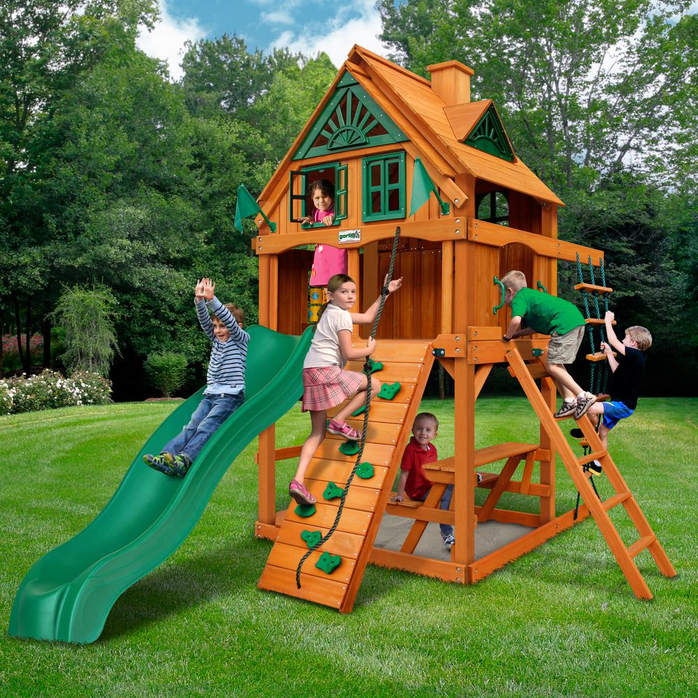 Cedar wooden playset with green wave slide, rock climbing wall, rope ladder, sandbox, and upper-level playhouse with shutters.