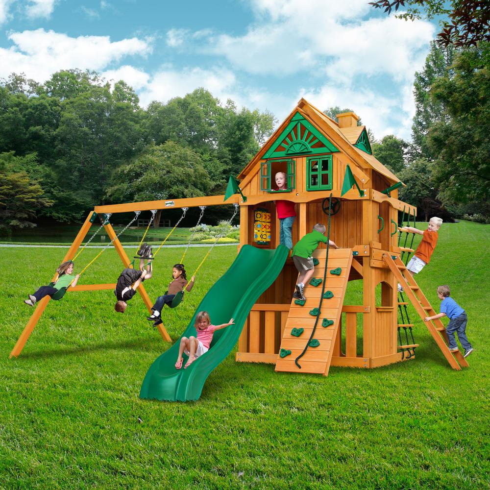 Cedar wooden playset with green slide, two belt swings, trapeze swing, climbing wall, rope ladder, and upper clubhouse with windows and shutters