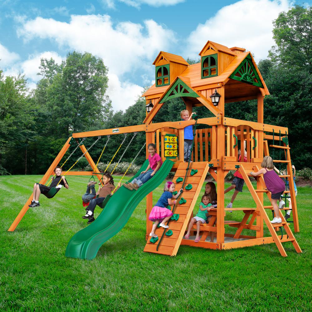 Cedar wooden playset with amber finish, green slide, swings, climbing wall, ladder, and playhouse roof with dormer windows.