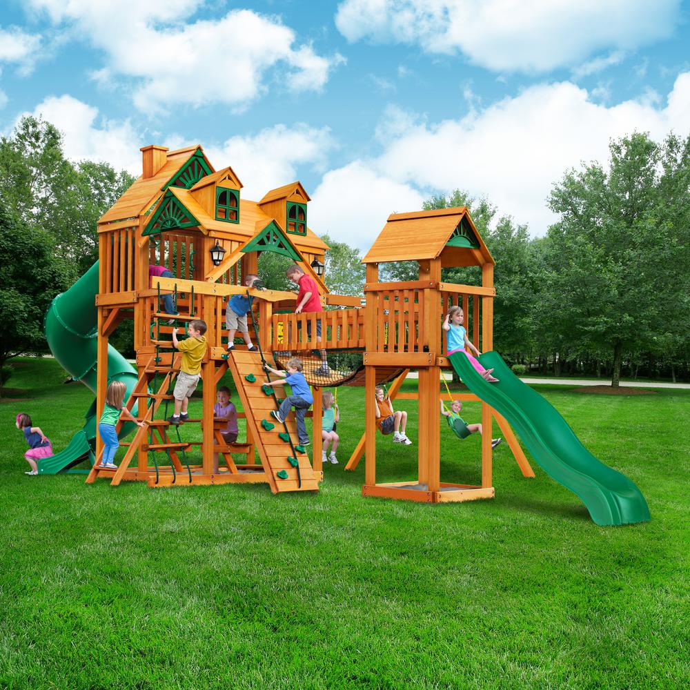 Wooden playset with green slides, climbing walls, swings, bridge, and children playing on grass under blue sky