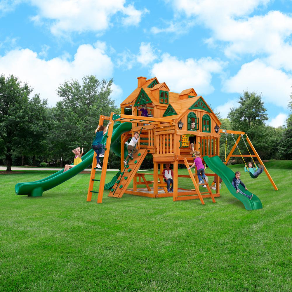 Cedar wooden playset with three green slides, swings, climbing wall, monkey bars, and multi-level decks with decorative roofs