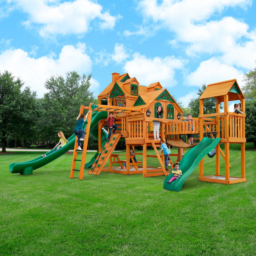 Wooden playset with two forts, green slides, climbing wall, monkey bars, tic-tac-toe board, and children playing outdoors