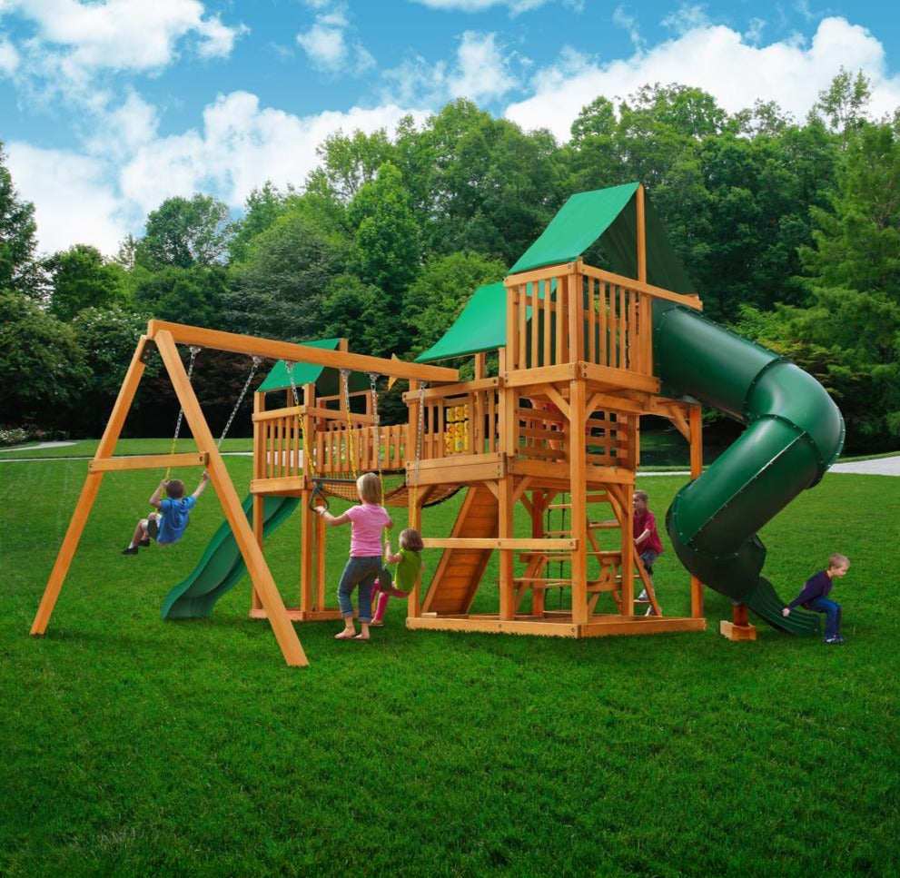 Wooden playset with green vinyl canopies, turbo tube slide, swings, climbing ladder, and rope ladder on grass lawn with children playing