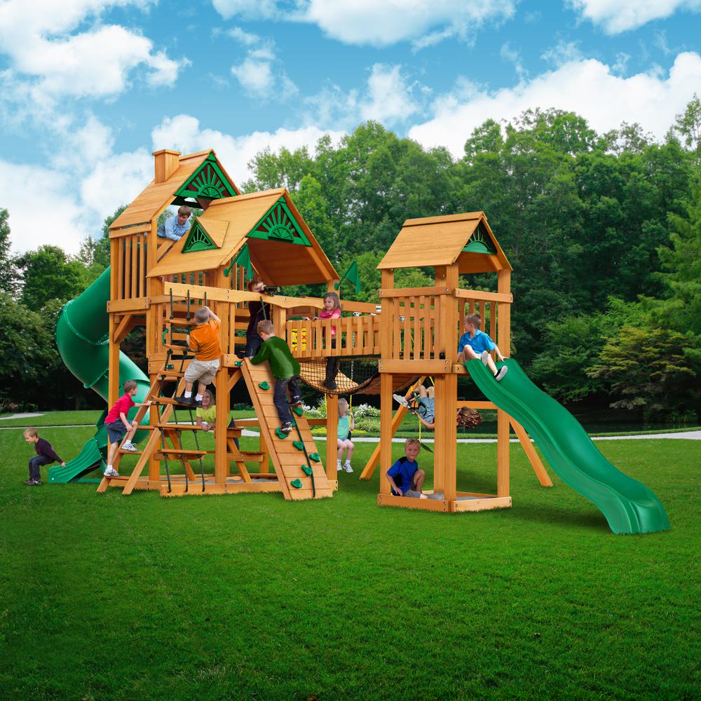 Cedar wooden playset with green slides, climbing walls, rope ladders, multiple towers, and children playing on grass